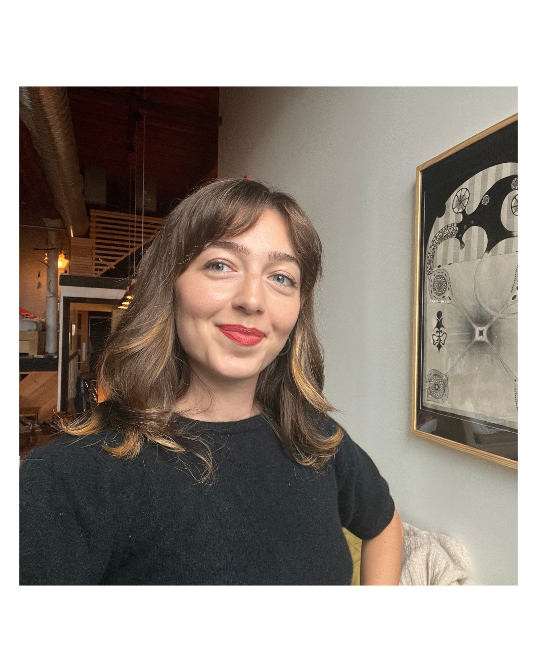 A photo of Ajda, wearing a black shirt, smiling at the camera. An art print is on the wall beside Ajda.