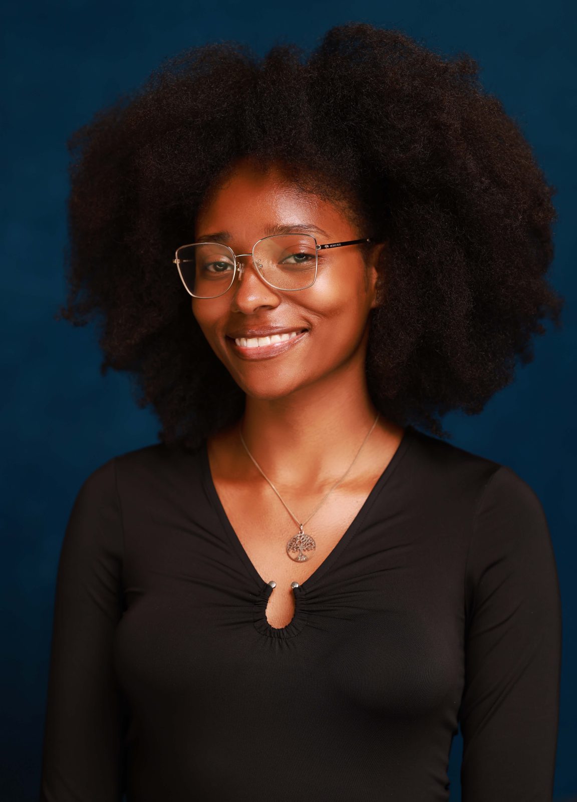 A photo of Kaitlyn against a professional blue background, wearing a black blouse and glasses, smiling at the camera.